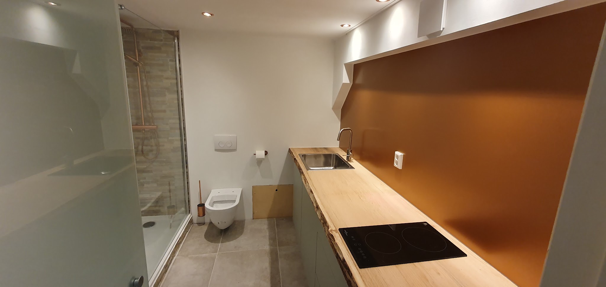 Compacte badkamer met keukenblok: inloopdouche, hangend toilet en houten werkblad met kookplaat tegen een koperkleurige wand